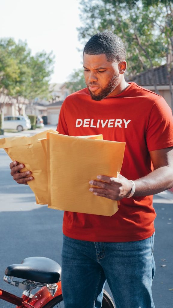 A delivery man in a red uniform holds envelopes while standing outdoors.