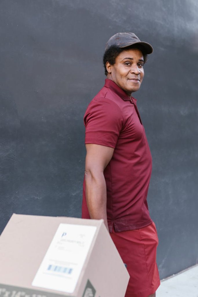 Smiling deliveryman in maroon uniform holding a box against a dark wall.